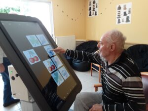 Résident de l’EHPAD utilisant une tablette tactile XXL pour un jeu de stimulation cognitive et de mémoire.